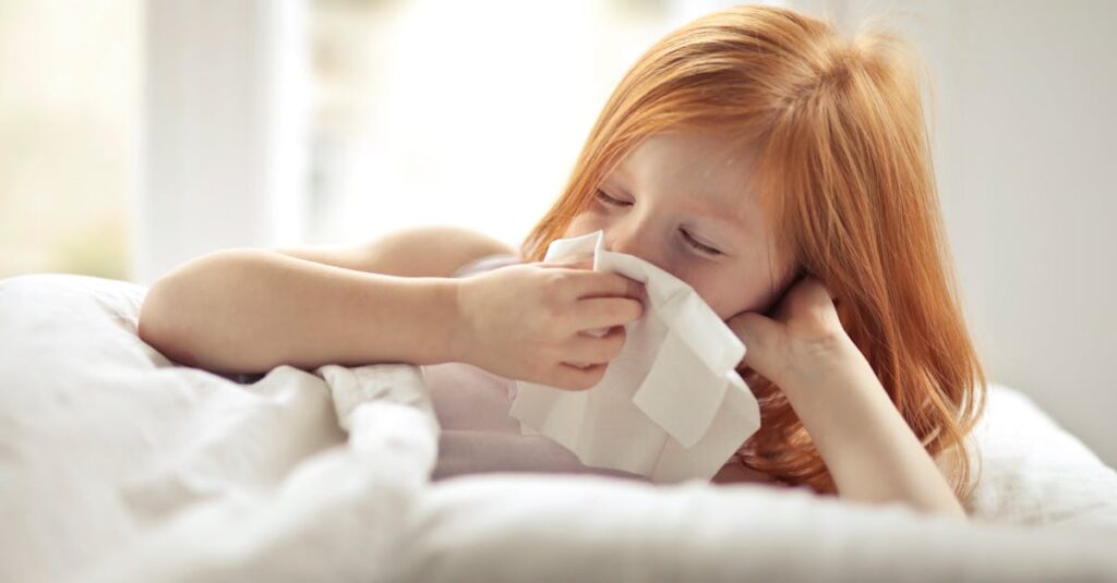 Redheaded child resting in bed, wiping nose with tissue, unwell with flu-like symptoms.