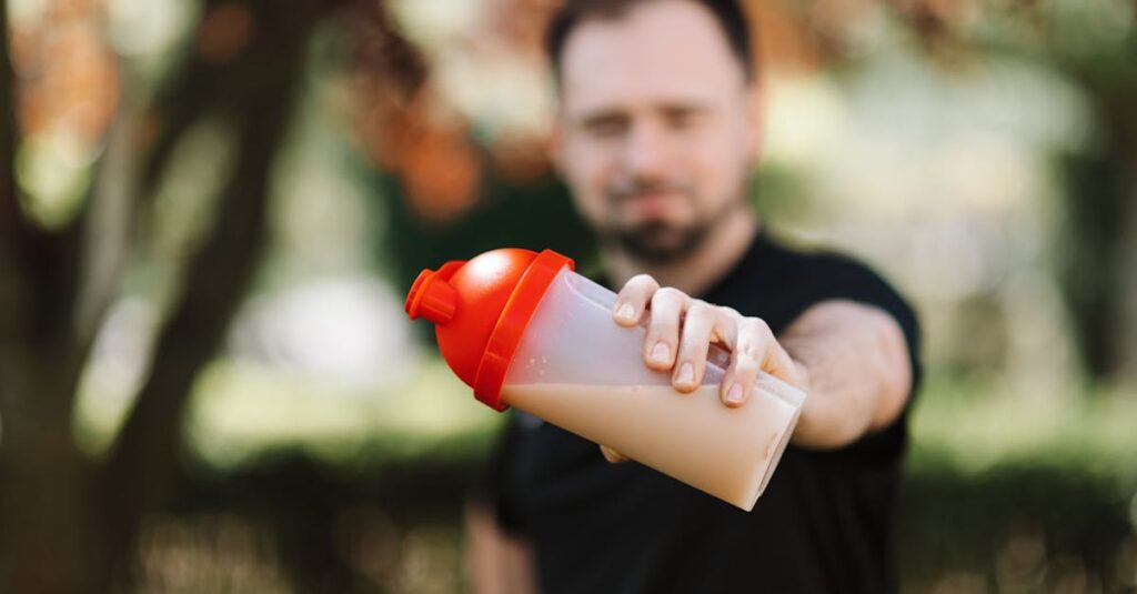 Man presenting a protein shake in a plastic tumbler against a blurred nature background.